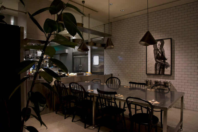 A dimly lit restaurant dining area next to the kitchen at TRUNK(HOTEL) CAT STREET features a metal table, black chairs, pendant lights, and a framed black-and-white portrait on a white tiled wall. A plant is partially visible in the foreground.