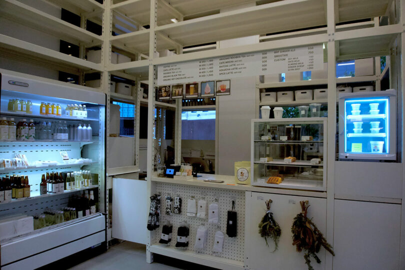A minimalist store interior at TRUNK(HOTEL) CAT STREET features white shelving, a refrigerator stocked with beverages, a counter with a menu above, and various packaged goods and dried herbs on display