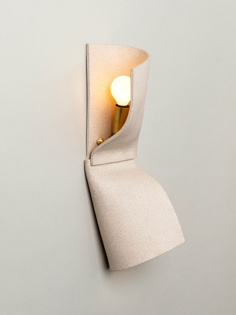 A wall-mounted Virignia Sin lighting fixture featuring a beige, folded, textured cover that partially reveals a round light bulb on a brass base