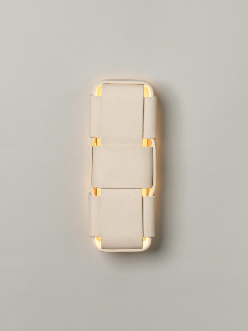 A modern wall sconce features a rectangular, layered design in neutral beige, emitting a soft, warm glow against a plain gray background