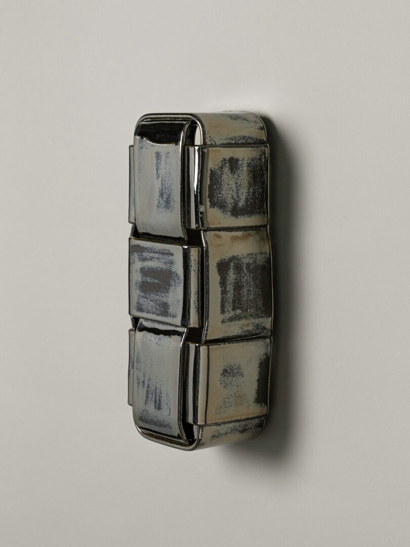 A metallic, rectangular object with six segments and a slightly worn, brushed finish—reminiscent of Virignia Sin lighting—is mounted vertically on a plain light surface