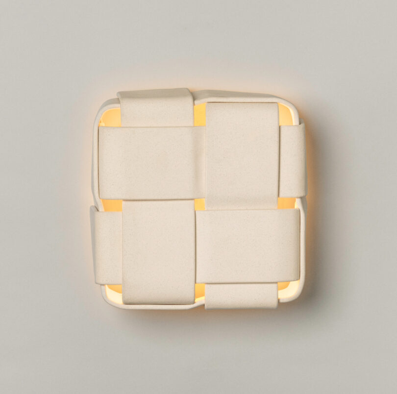 Rectangular wall sconce with interwoven white ceramic bands, part of the Virginia Sin lighting collection, casts a warm glow softly from behind against a neutral background