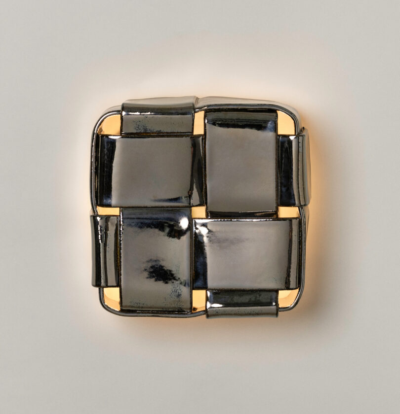 A square metallic wall sconce with a woven pattern, inspired by Virginia Sin lighting, emits a warm light from behind and is mounted on a plain light-colored wall