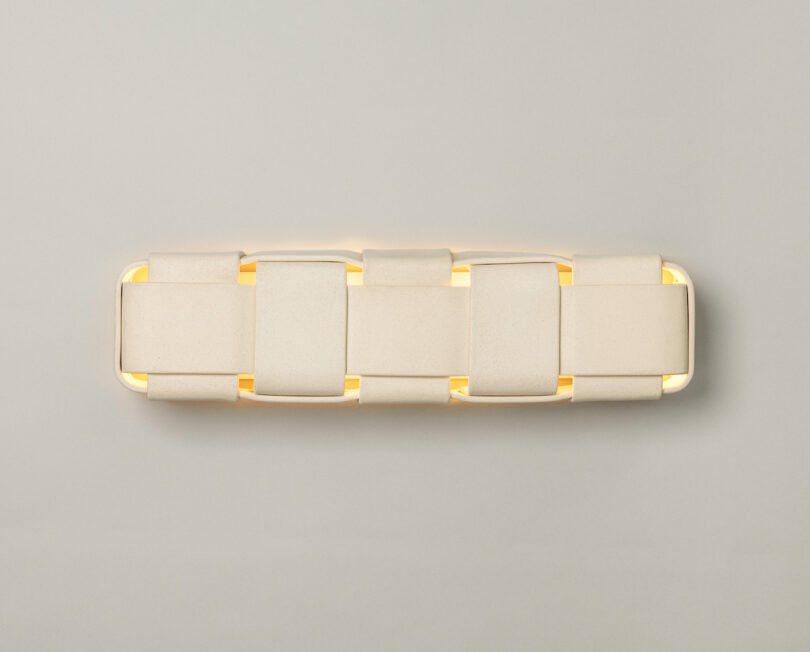 Rectangular wall light fixture with a woven, geometric design emits a soft, warm glow against a plain light gray background, capturing the signature artistry of Virginia Sin lighting