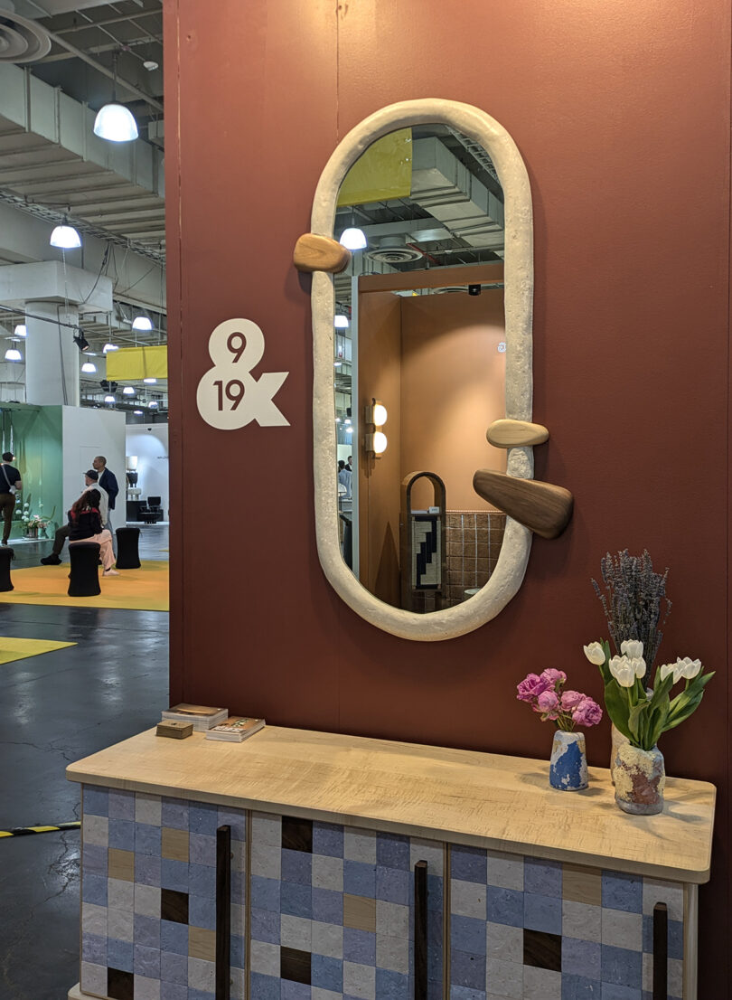 A large oval mirror with irregular stone accents hangs on a reddish-brown wall above a mosaic-patterned sideboard with flowers and books, at a trade show booth labeled "9 & 19".