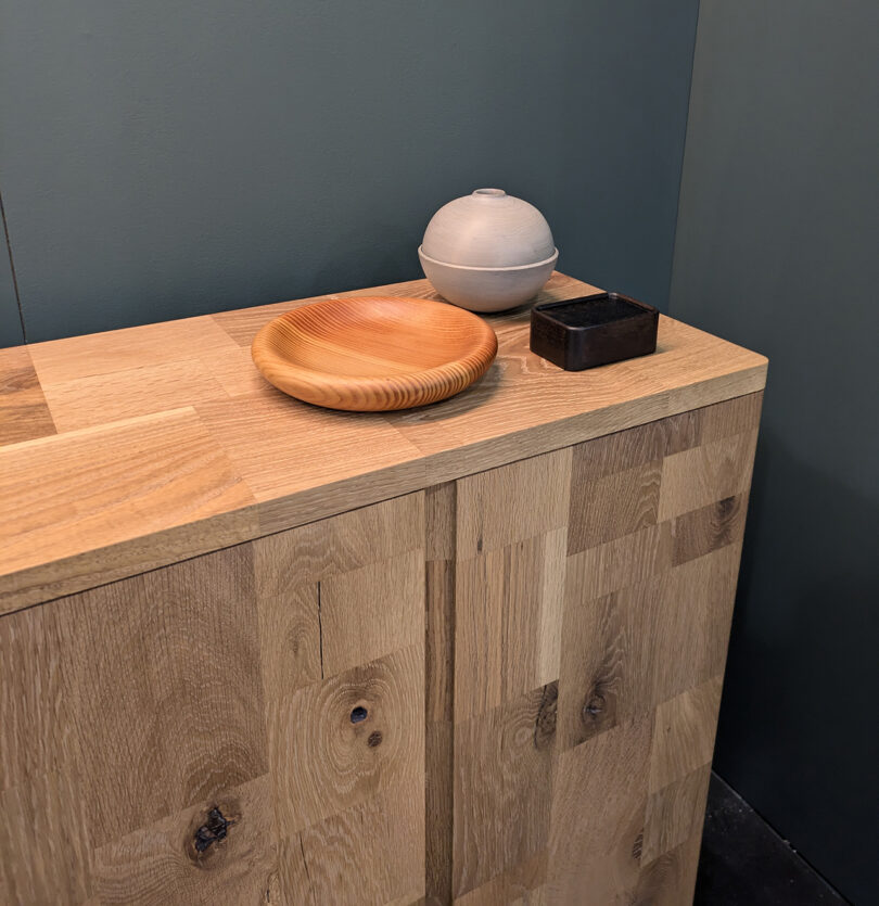 A wooden cabinet with a round wooden plate, a gray spherical container, and a small rectangular black box on top against a green wall.