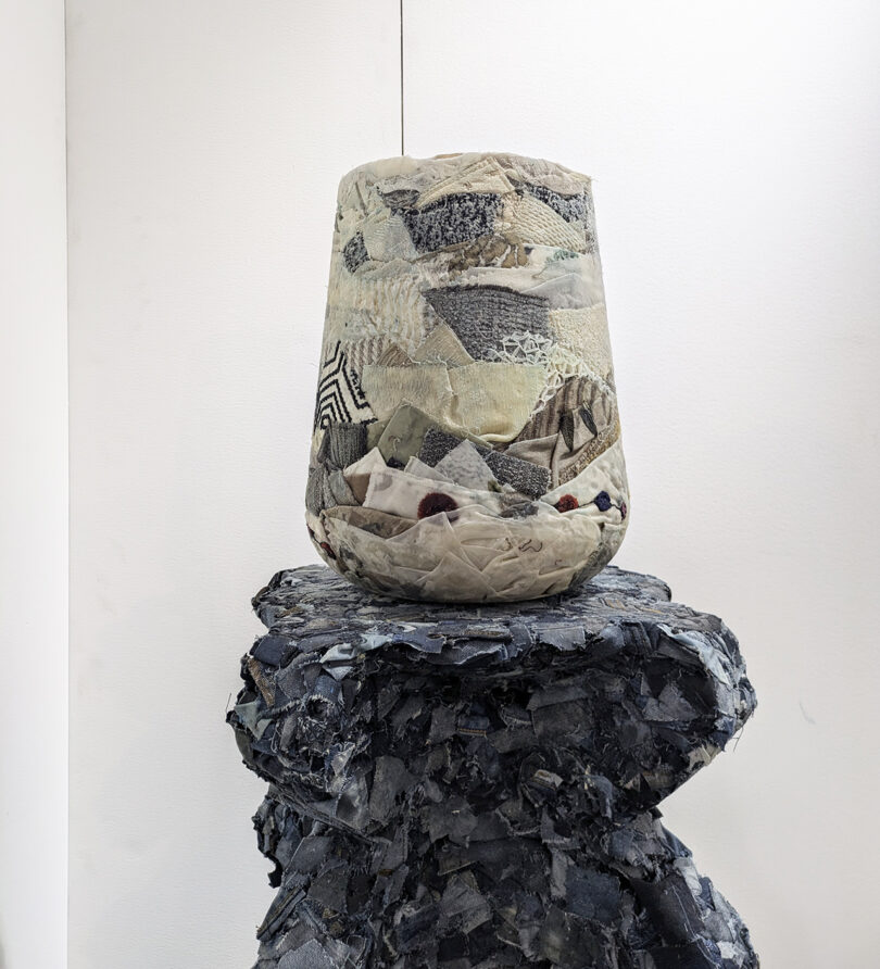 A textured ceramic vase with layered, patterned surfaces sits on a rough, dark stone pedestal against a plain white background.