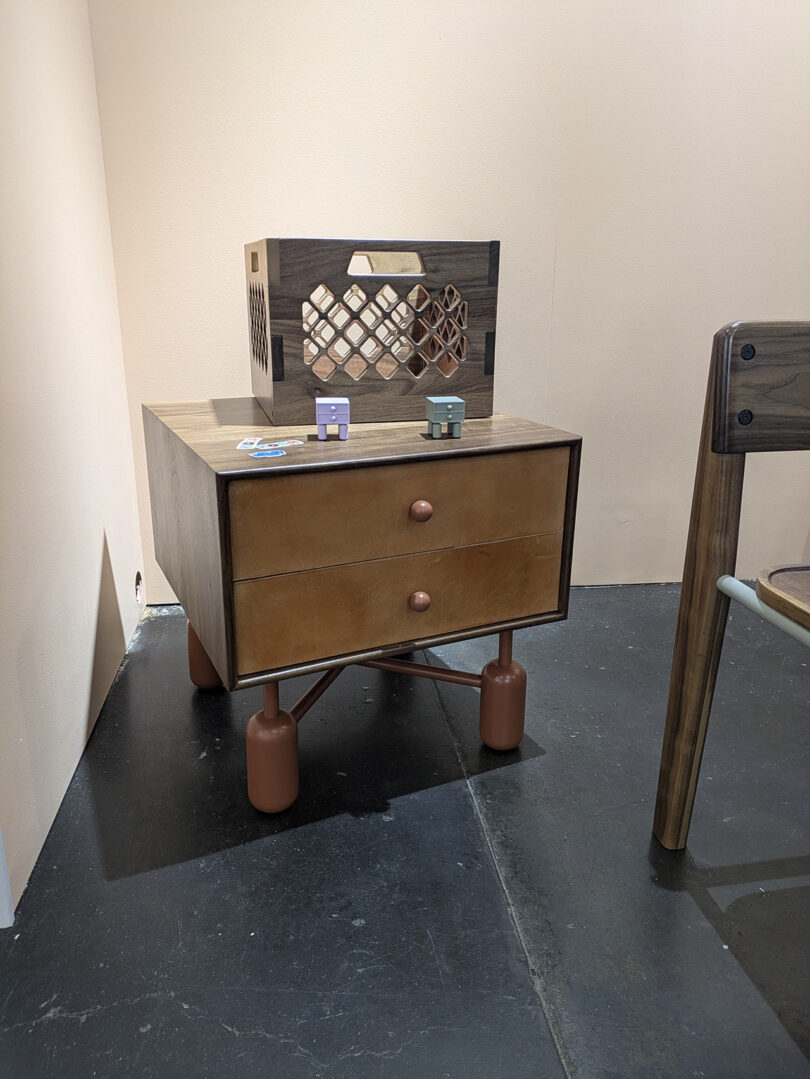 A modern wooden nightstand with two drawers and round legs, topped with small block toys and a wooden crate, stands on a dark floor against beige walls.