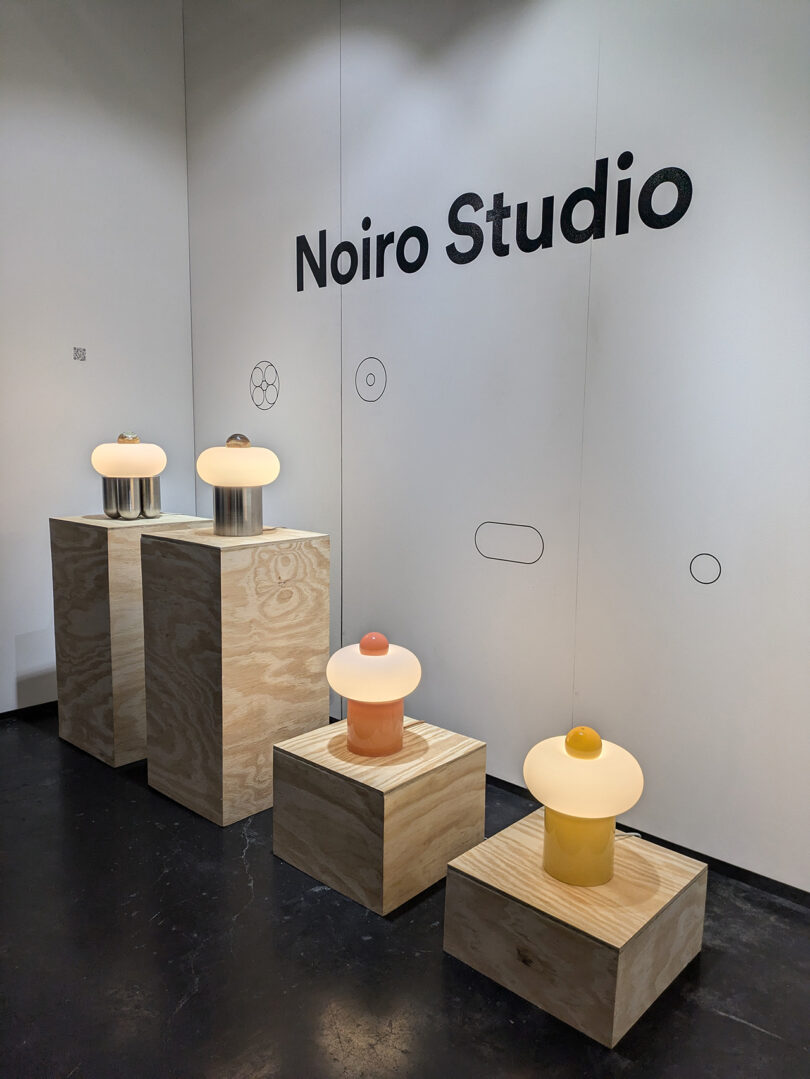 Four modern table lamps are displayed on wooden plinths in a gallery space with "Noiro Studio" written on the white wall behind them.