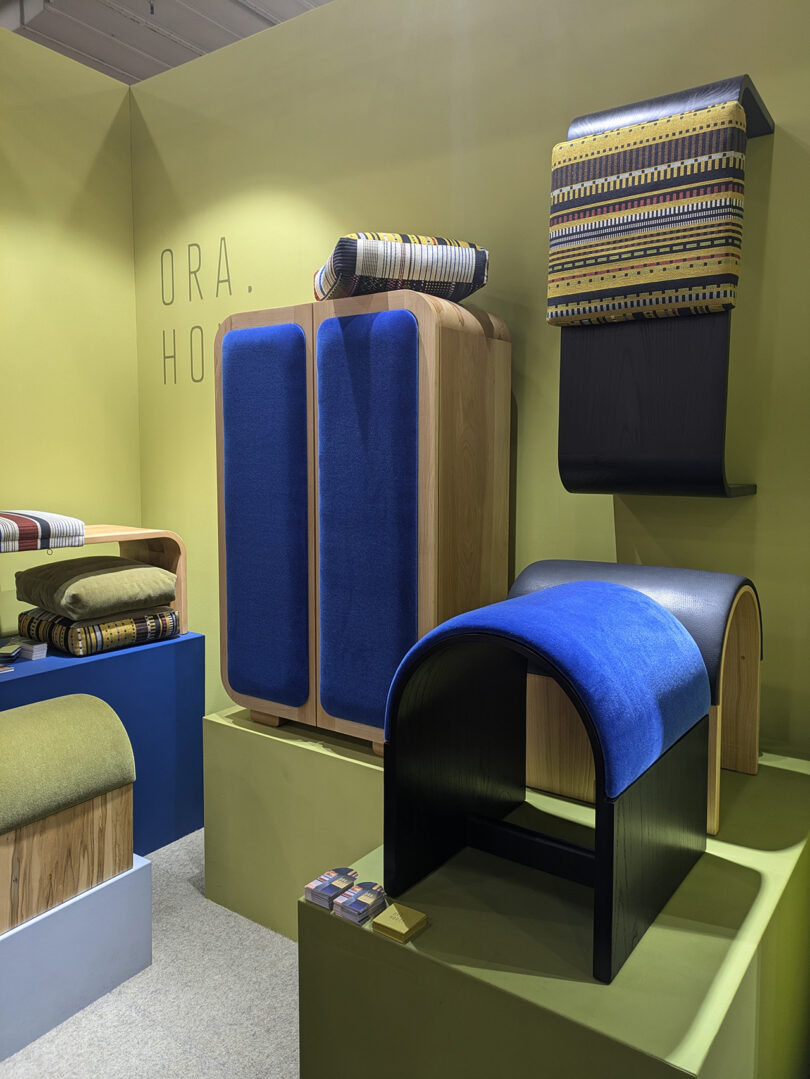 Display of modern furniture pieces with colorful cushions and textiles arranged on platforms against a yellow wall with "ORA." and "HO." text.