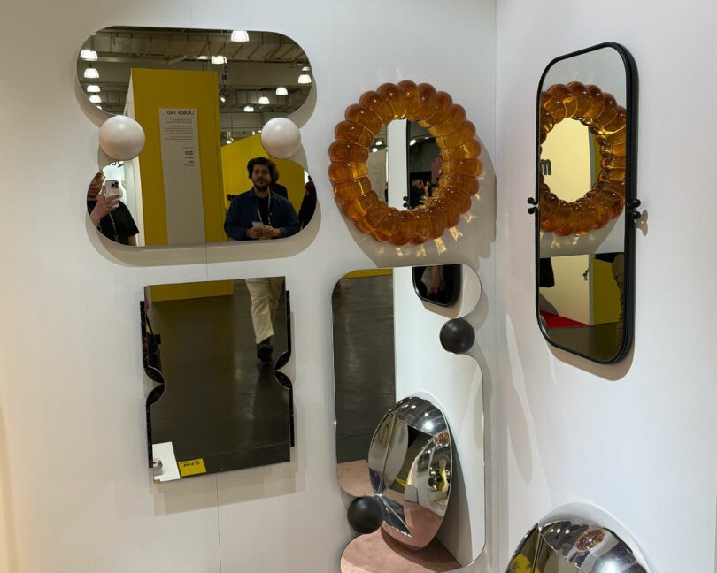 A display of various uniquely shaped mirrors on a white wall at Wanted Design 2025, reflecting people and the exhibition hall in the background.