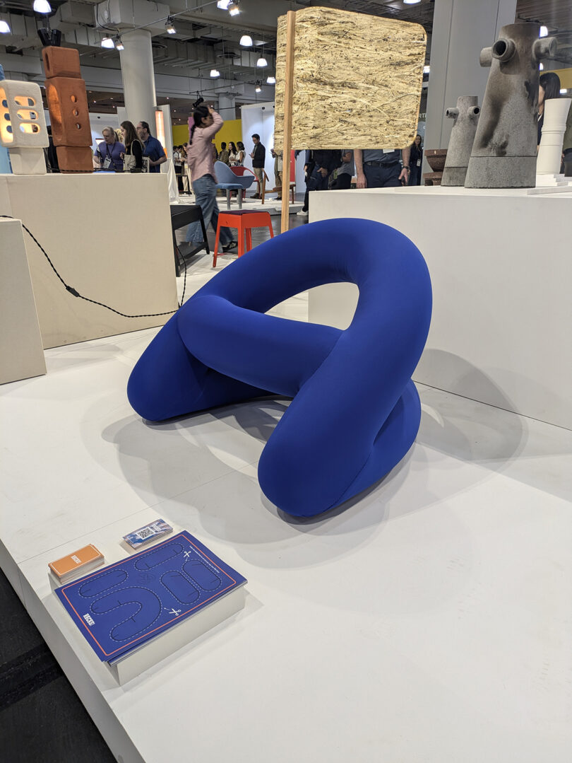 A blue, abstract, tubular chair sits on display in an exhibition space surrounded by other modern art and design pieces.
