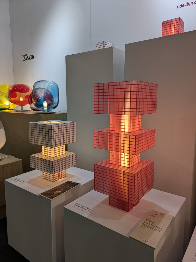 Three geometric lamps with grid-patterned, translucent cubes are displayed on white pedestals in an exhibition space, with informational sheets placed in front of them.