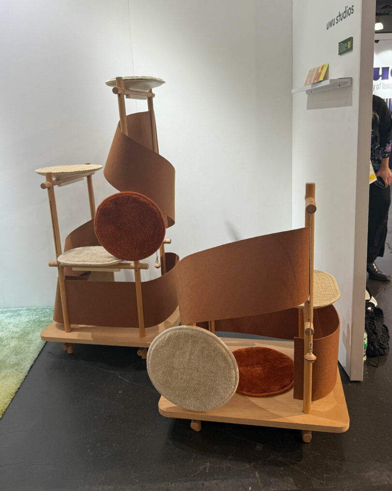 Two wooden cat trees with circular platforms, brown and beige cushions, and a winding brown ramp are displayed in an indoor exhibition setting at Wanted Design 2025.