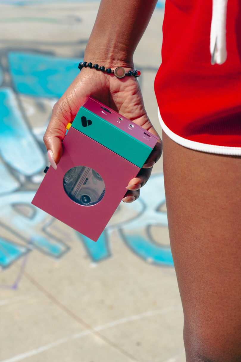 A person in red athletic shorts holds a colorful, boxy WE-001 Edith camera with a circular lens, standing on pavement with blue graffiti in the background.