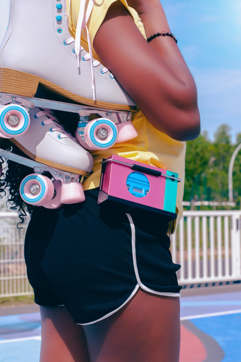 Person in black shorts and yellow top holding a pair of white and pink roller skates over their shoulder, with a colorful camera clipped to their waist—WE-001 Edith style—outdoors on a sunny day.