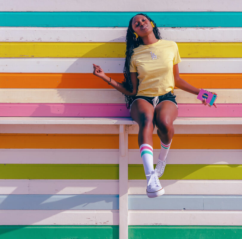 WE-001 Edith, dressed in a yellow shirt and shorts, sits on a bench against a vibrant, multicolored striped wall, listening to music with headphones and holding her phone.