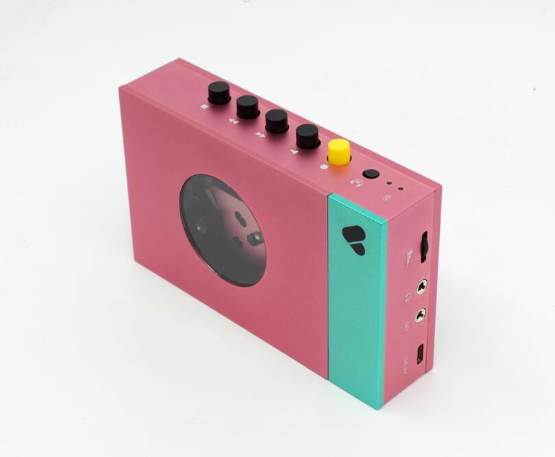 Rectangular pink and teal WE-001 Edith cassette player with black and yellow buttons on top, a transparent window, and side ports, placed on a white background.