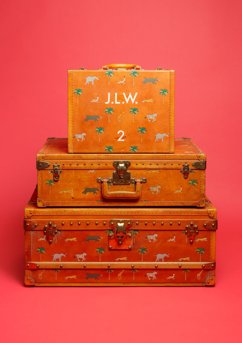 Three stacked vintage-style orange suitcases with animal and palm tree patterns on a red background; top suitcase labeled "J.L.W. 2.