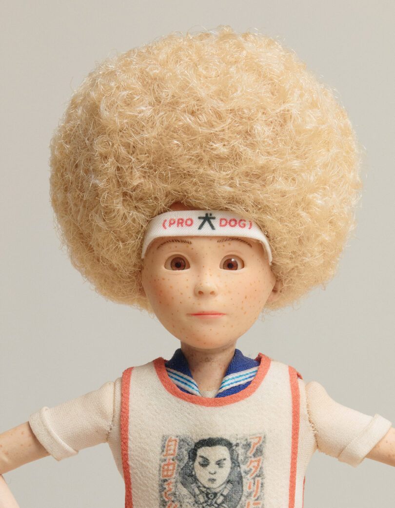 A doll with a large blonde afro, freckled face, wearing a headband labeled “PRO DOG,” and a shirt featuring a portrait and Japanese text.