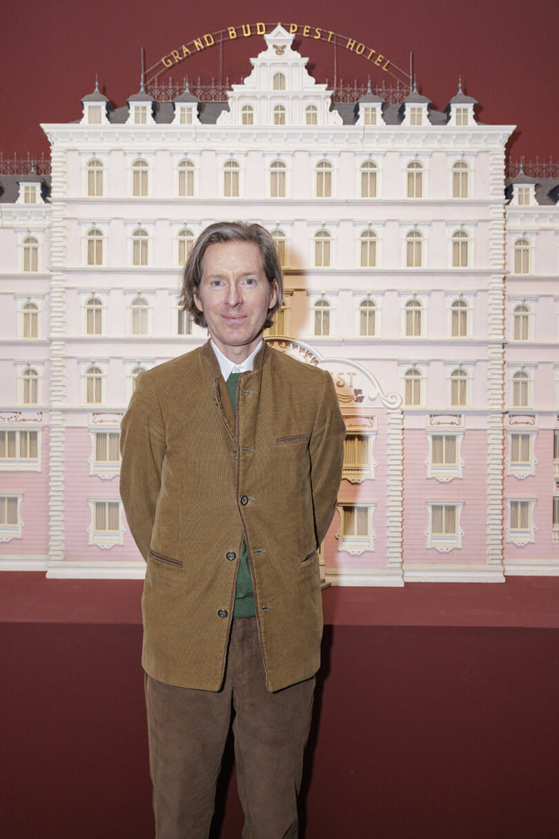 A man in a brown corduroy suit stands in front of a model of the Grand Budapest Hotel with a red background.