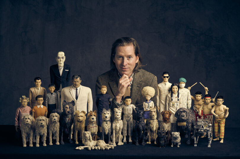 A man sits behind an array of dog and human figurines arranged in rows against a dark background.