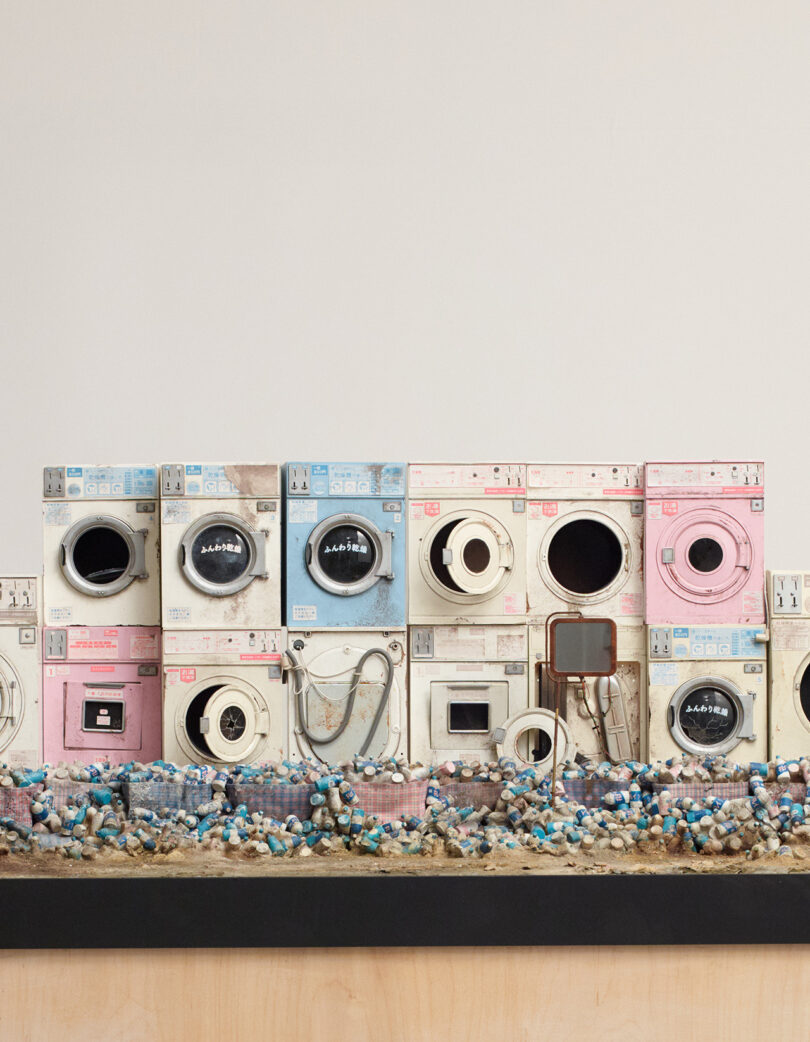 A row of old, weathered washing machines and dryers is displayed above a base covered with scattered debris and tangled wires.
