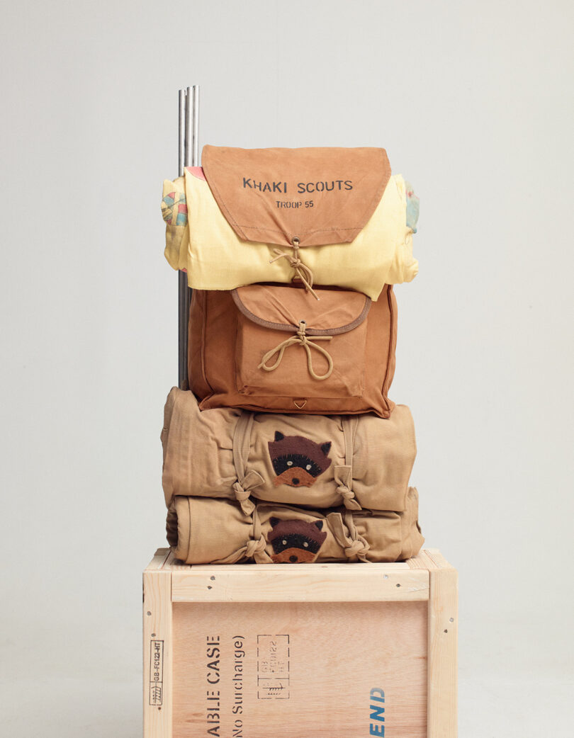 A tan backpack labeled "KHAKI SCOUTS Troop 55" stacked on rolled blankets and a crate, with patches visible on the blankets.