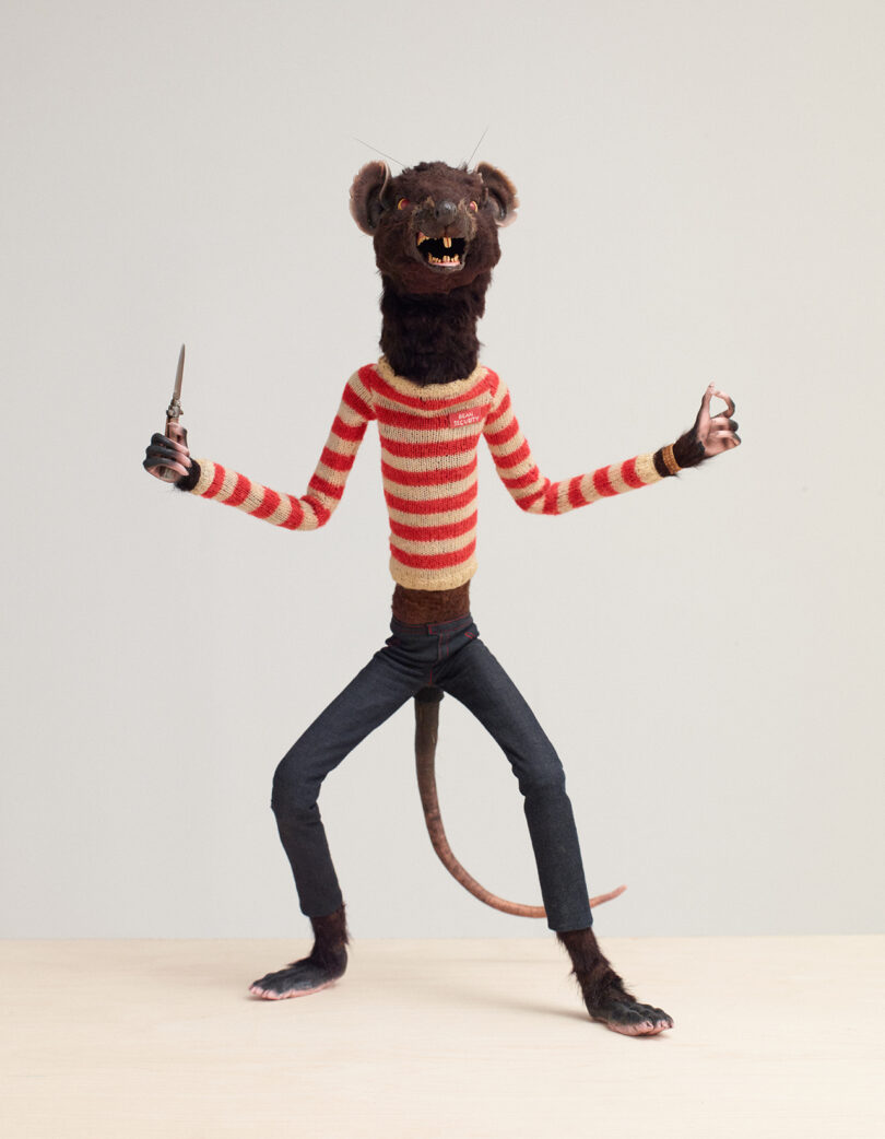 A humanoid rat figure with a striped sweater, jeans, and a tail poses holding a knife against a plain background.