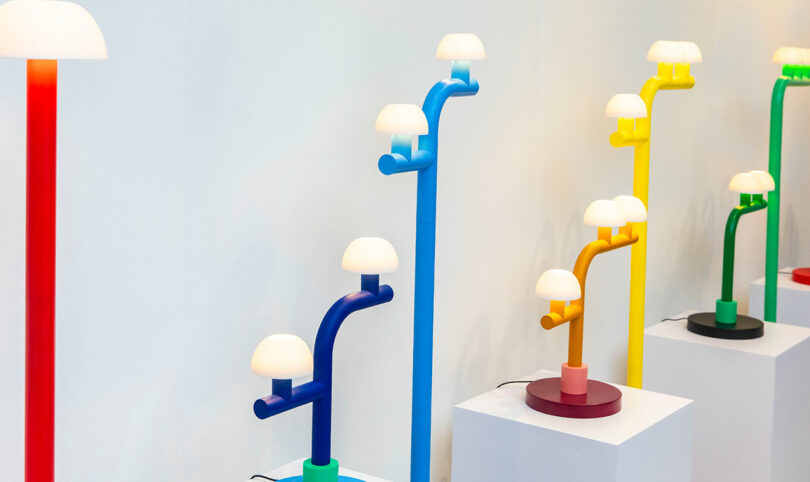 A display of colorful, modern lamps by Nader Gammas in red, blue, purple, yellow, and green, each with multiple white dome-shaped lights on white pedestals against a plain wall.