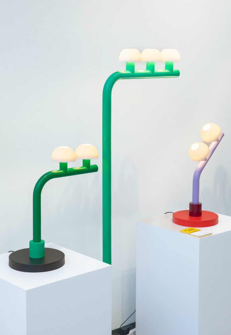 Two modern table lamps by Nader Gammas, featuring curved stems and multiple dome-shaped bulbs, are displayed on white pedestals against a plain white wall.