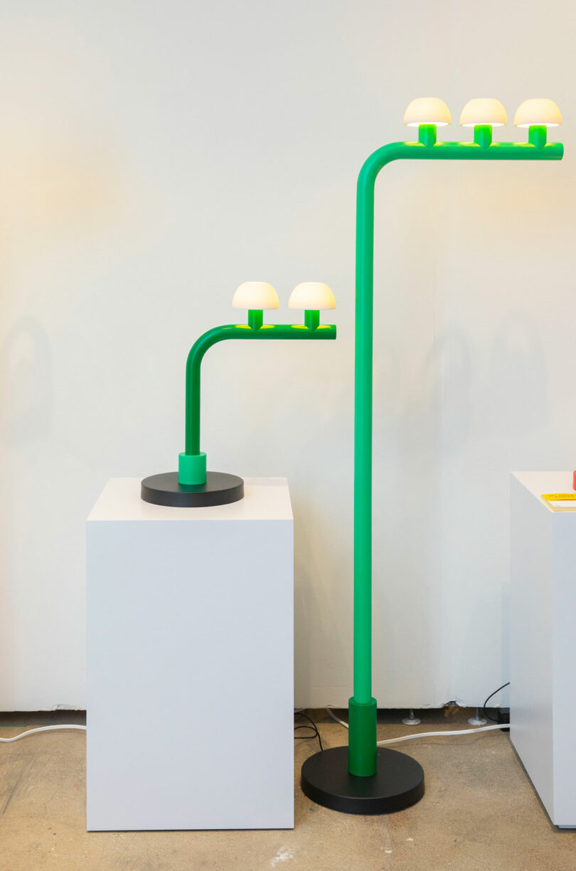 A green lamp and a matching green floor lamp by Nader Gammas, each with multiple white bulb covers, are displayed on white pedestals against a plain wall.