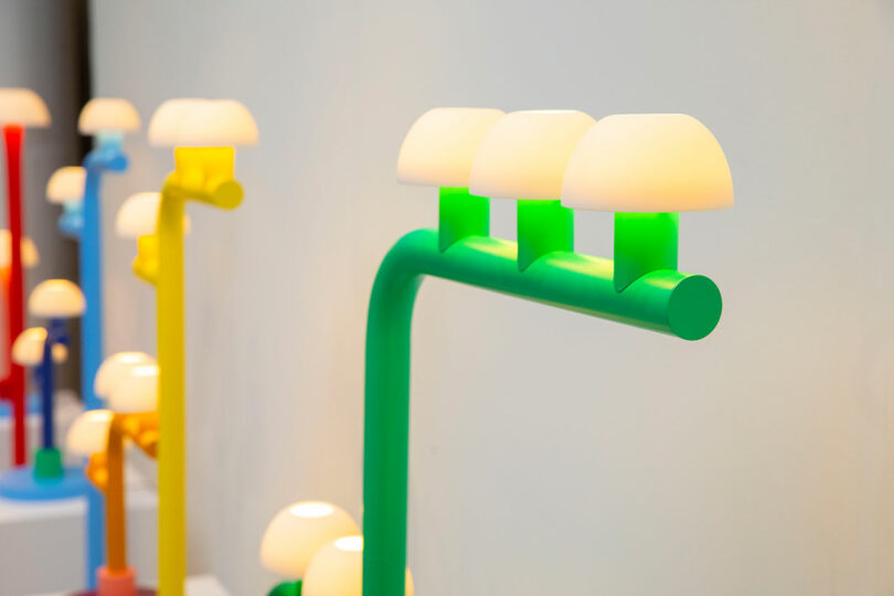 Colorful modern lamps by Nader Gammas feature rounded white shades and vibrant yellow, green, and blue bases, displayed against a plain, light-colored background.