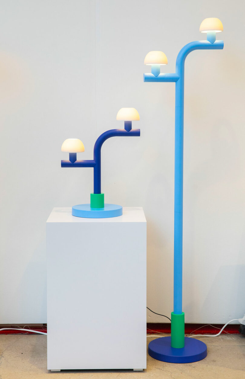 Two modern, blue lamps with multiple branches and dome-shaped bulbs, designed by Nader Gammas—one perched on a white pedestal, the other standing on the floor—are set against a plain wall.