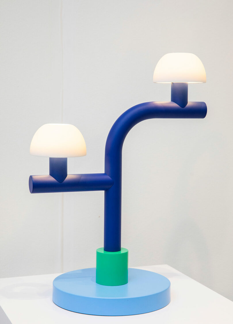 A modern table lamp by Nader Gammas, featuring a blue, branch-like structure, two white dome lights, and a round blue base with a green accent, elegantly displayed on a white surface.
