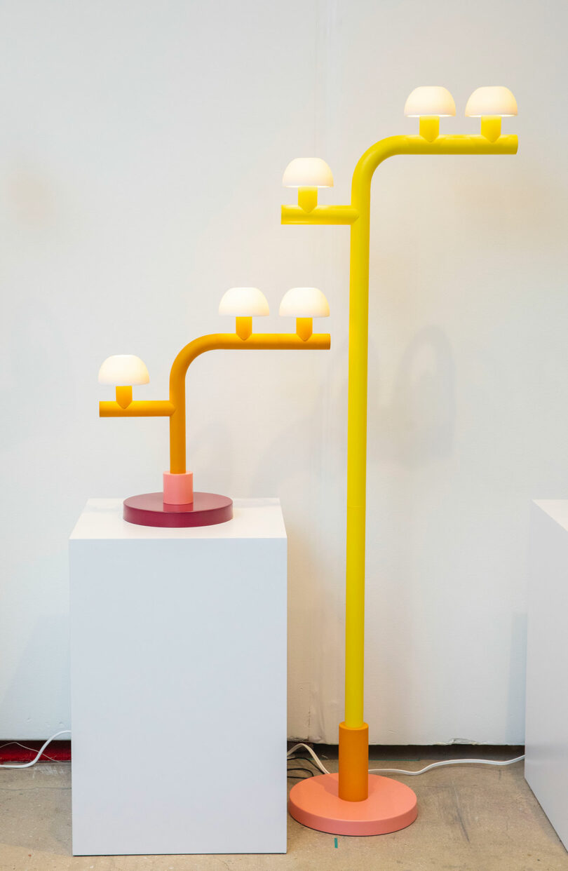 Two modern lamps by Nader Gammas, featuring yellow stems and multiple small white dome-shaped shades, are displayed on separate white pedestals against a plain wall.