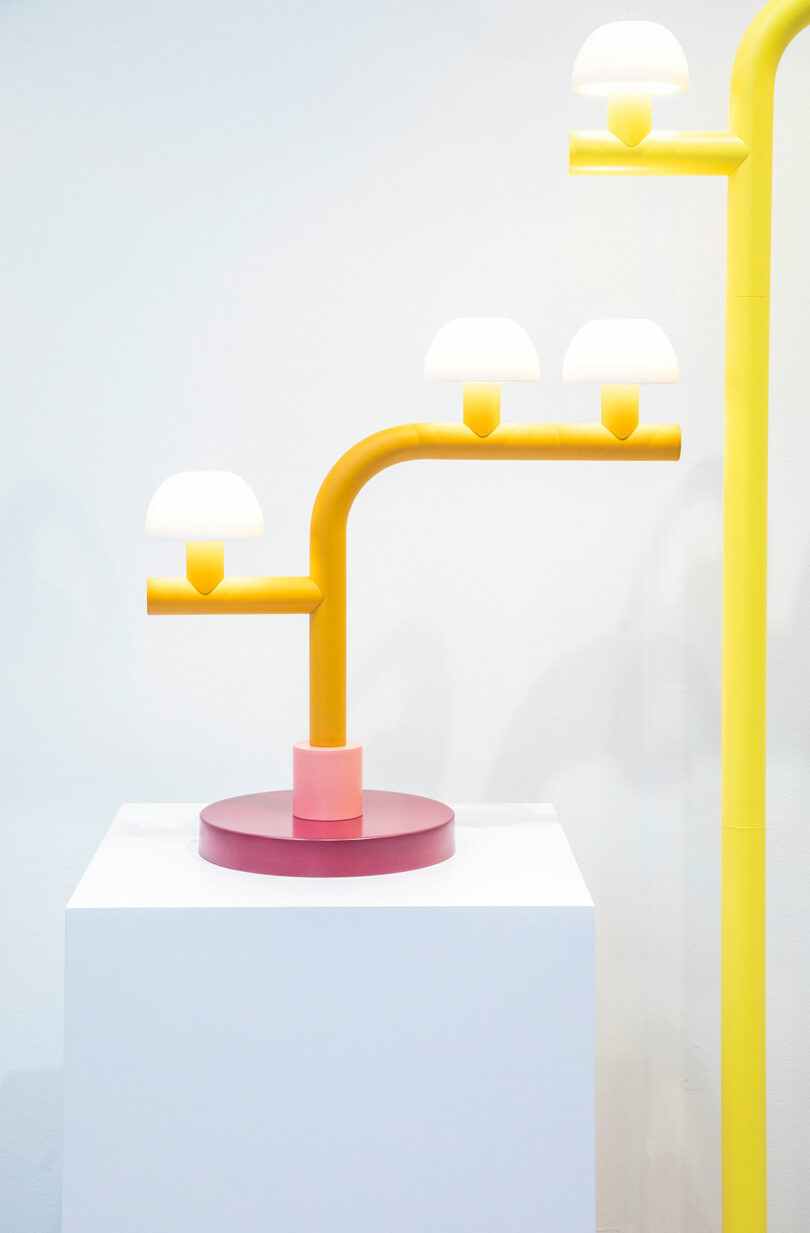 A modern lamp by Nader Gammas features a pink base and yellow arms, each holding small dome-shaped white lights, elegantly displayed on a white pedestal against a light background.