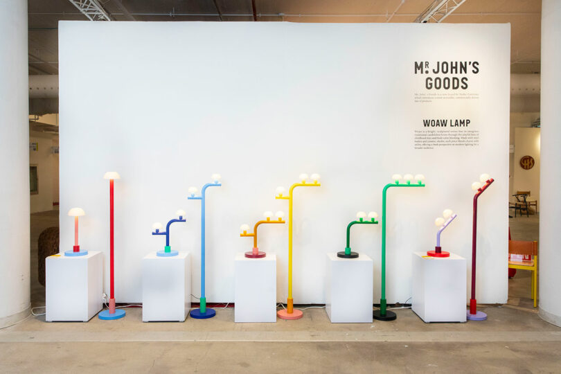 A display of colorful lamps in blue, yellow, green, and red—curated by Nader Gammas—are arranged on white pedestals in front of a wall with text reading 