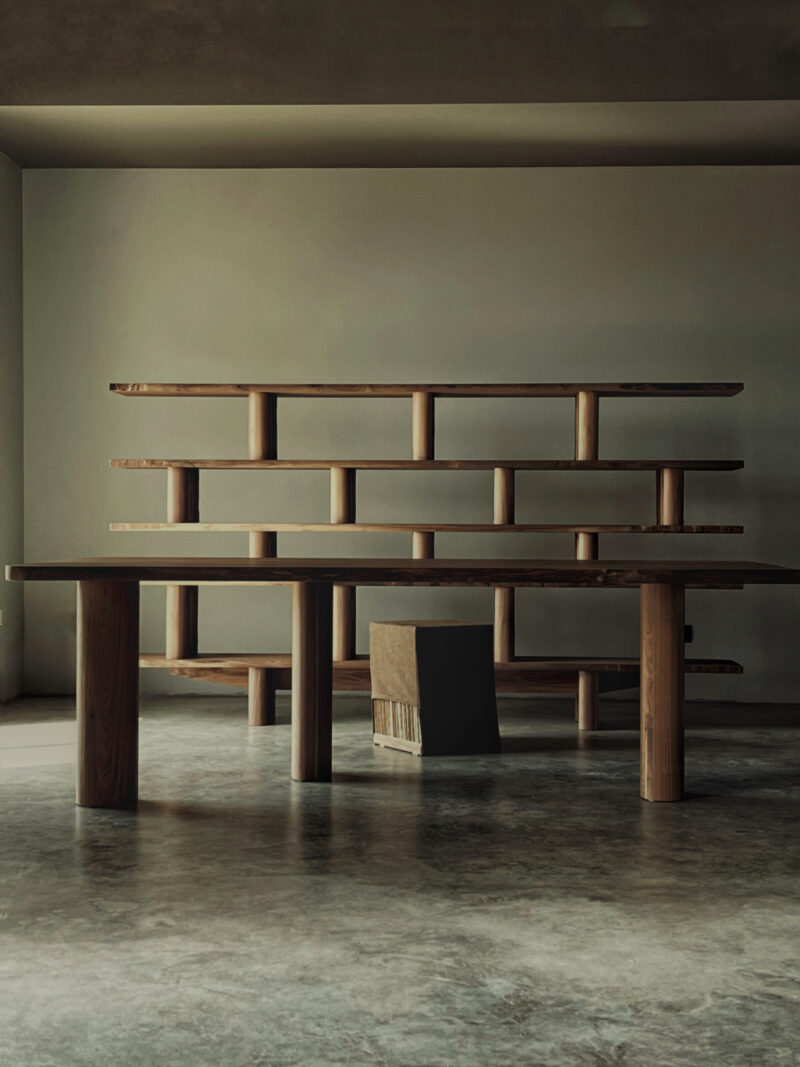 Sculptural wooden table and bookshelf in a minimal interior with a polished concrete floor.