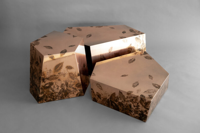 Three metallic, geometric tables with a reflective bronze finish and leaf-like patterns are arranged together on a plain gray background