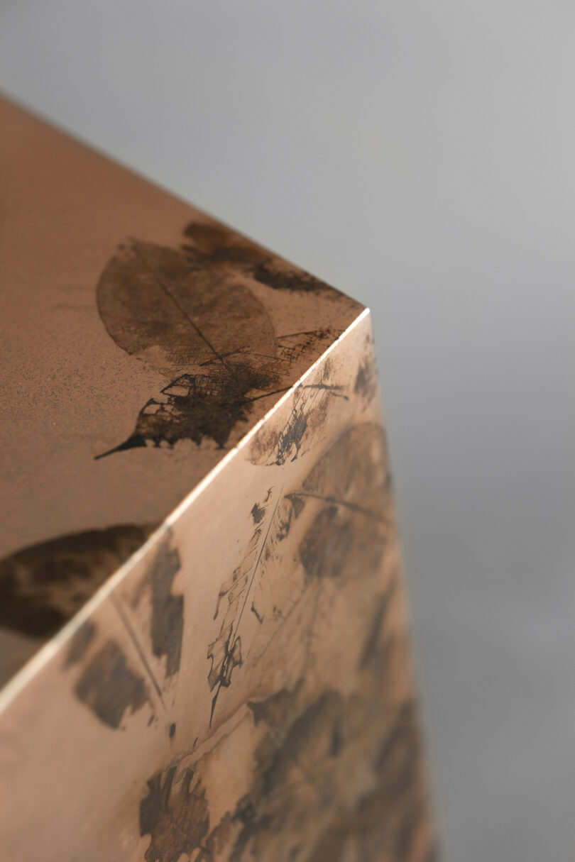 A close-up of a metallic copper-colored surface with leaf imprints, showing a sharp corner against a blurred gray background