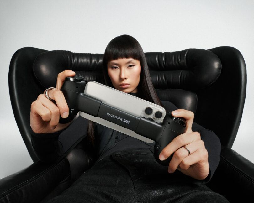 A person sitting in a black chair holds a smartphone gaming controller labeled "Backbone One" with a phone mounted in the device.