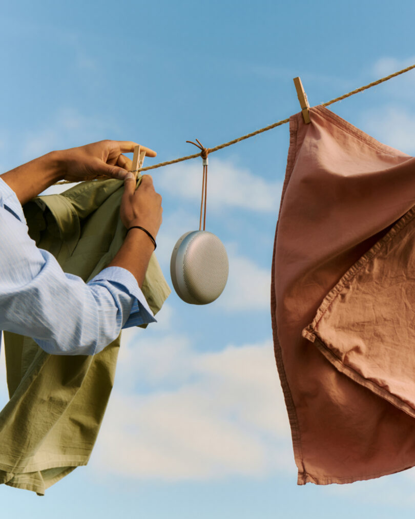 A person hangs green and pink fabric on a clothesline with clothespins beside a portable speaker under a blue sky.
