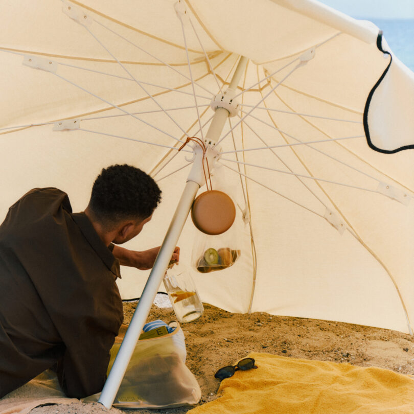 A person sits under a large beach umbrella on sand, reaching for a jar of water next to a yellow towel, sunglasses, and a bag.