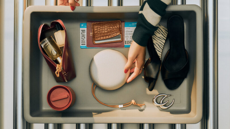 A plastic tray at airport security holds a passport, boarding pass, wallet, shoes, jewelry, travel mug, zippered pouch, and a portable speaker; a hand reaches for the speaker.