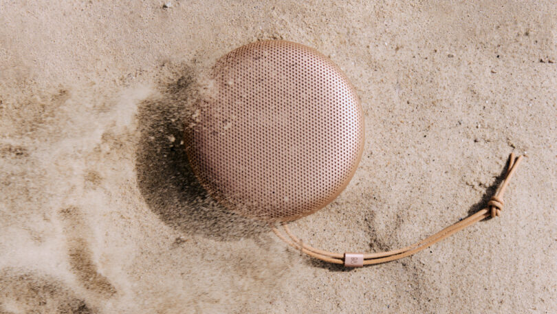 A round, portable speaker with a leather strap lies on sandy ground, partially covered by sand.