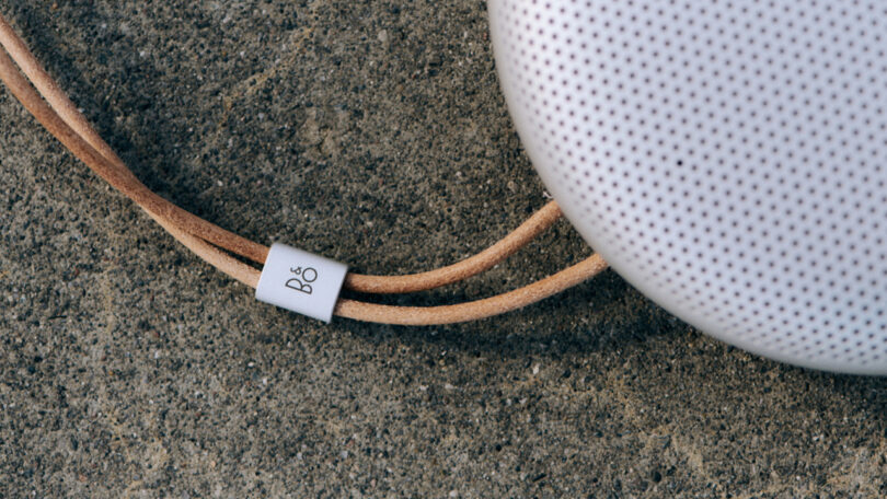 Close-up of a portable speaker with a perforated surface and a tan leather strap on a concrete background.