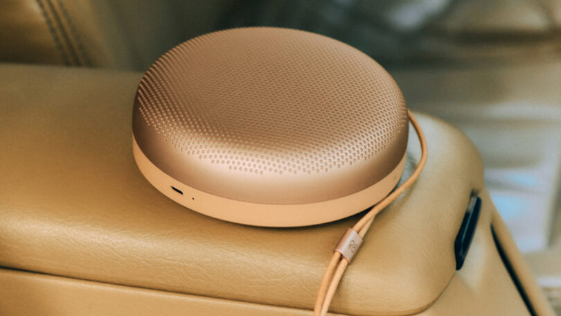 A round, gold-colored portable Bluetooth speaker with a textured top, resting on a tan leather car seat.