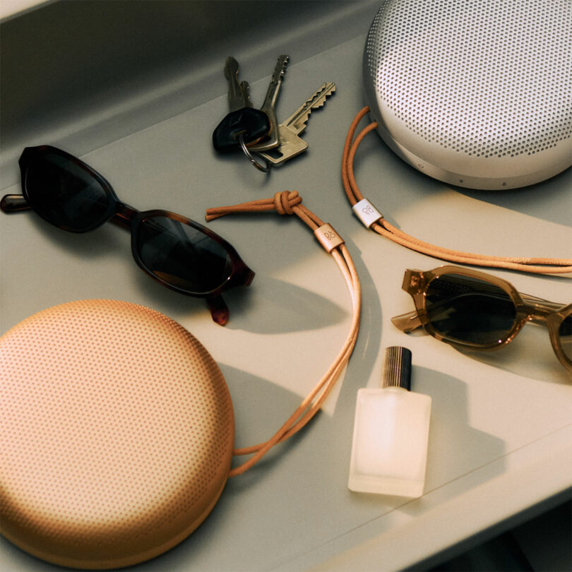 Two Beosound A1 portable speakers, two pairs of sunglasses, a set of keys, and a small bottle are arranged on a flat surface.