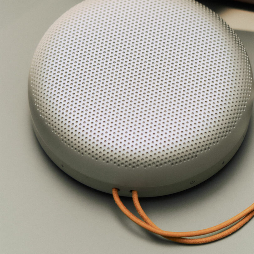 A round, portable Beosound A1 wireless speaker with a perforated metal grille and a tan wrist strap, placed on a flat surface.